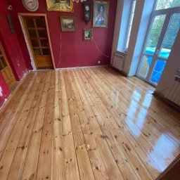 Grindelwald - Świeżo polakierowana drewniana podłoga w pokoju z czerwonymi ścianami, kryształowym żyrandolem i obrazami w złotych ramach.