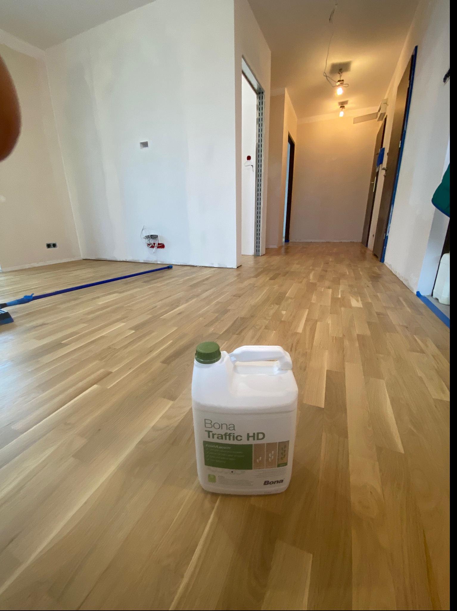 Świeżo polakierowana, jasna podłoga parkietowa z widocznym pojemnikiem Bona Traffic HD i fragmentem narzędzia do rozprowadzania lakieru, zabezpieczona taśmą malarską przy ścianach.