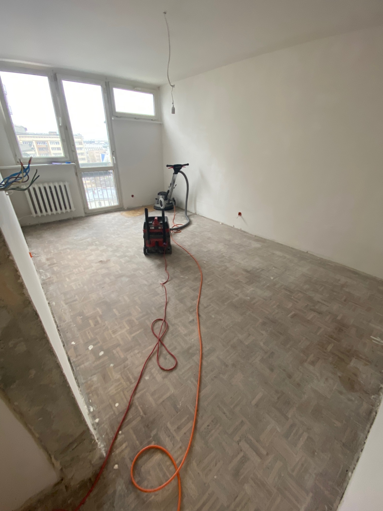 Pokój w trakcie remontu, z widocznym parkietem ułożonym w jodełkę, częściowo wyszlifowanym, z maszyną do szlifowania parkietu i odkurzaczem przemysłowym w tle, przy oknie widoczne kable elektryczne.