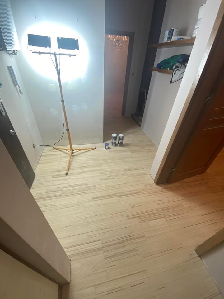 Wnętrze w trakcie remontu z nową, jasną podłogą z paneli, lampą roboczą na trójnogu i puszkami farby w tle.
