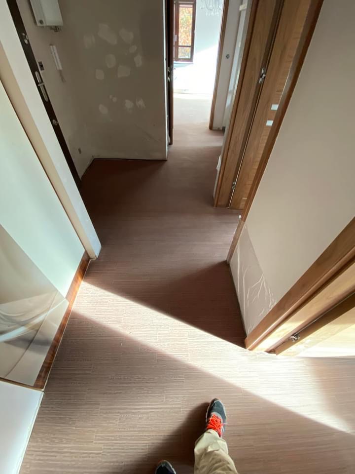 Korytarz z nową, brązową podłogą imitującą drewno, widok z góry na fragment butów i spodni osoby fotografującej, ściany częściowo przygotowane do malowania, widoczne drzwi z drewnianymi ościeżnicami.