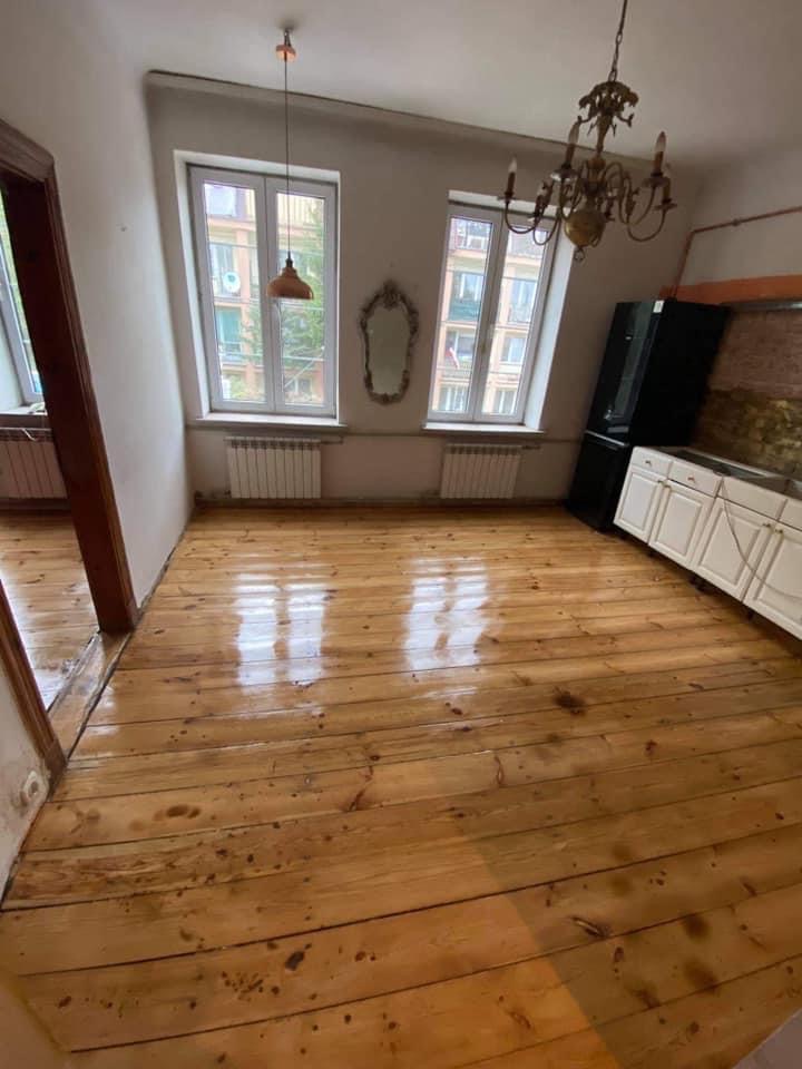 Wycyklinowana podłoga z desek w pokoju z dwoma oknami, widoczne ślady po poprzednich zabrudzeniach, sufit ze skosem i wiszącymi lampami.