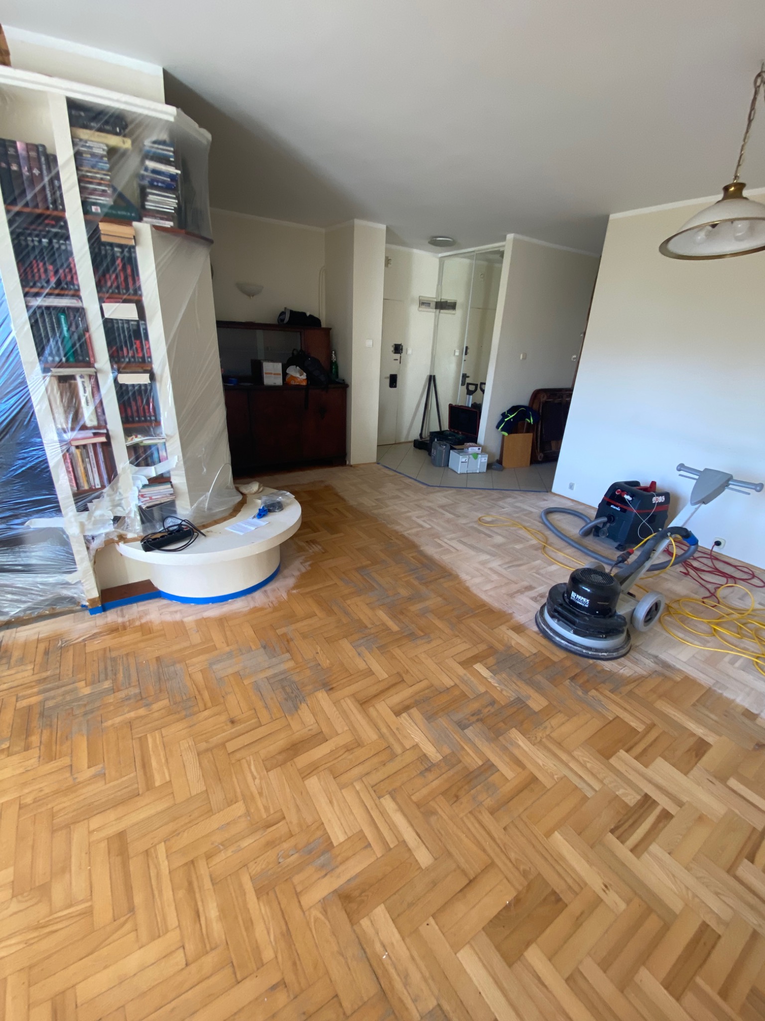 Parkiet w jodełkę częściowo wyszlifowany, z widoczną maszyną do szlifowania i odkurzaczem przemysłowym, meble zabezpieczone folią.