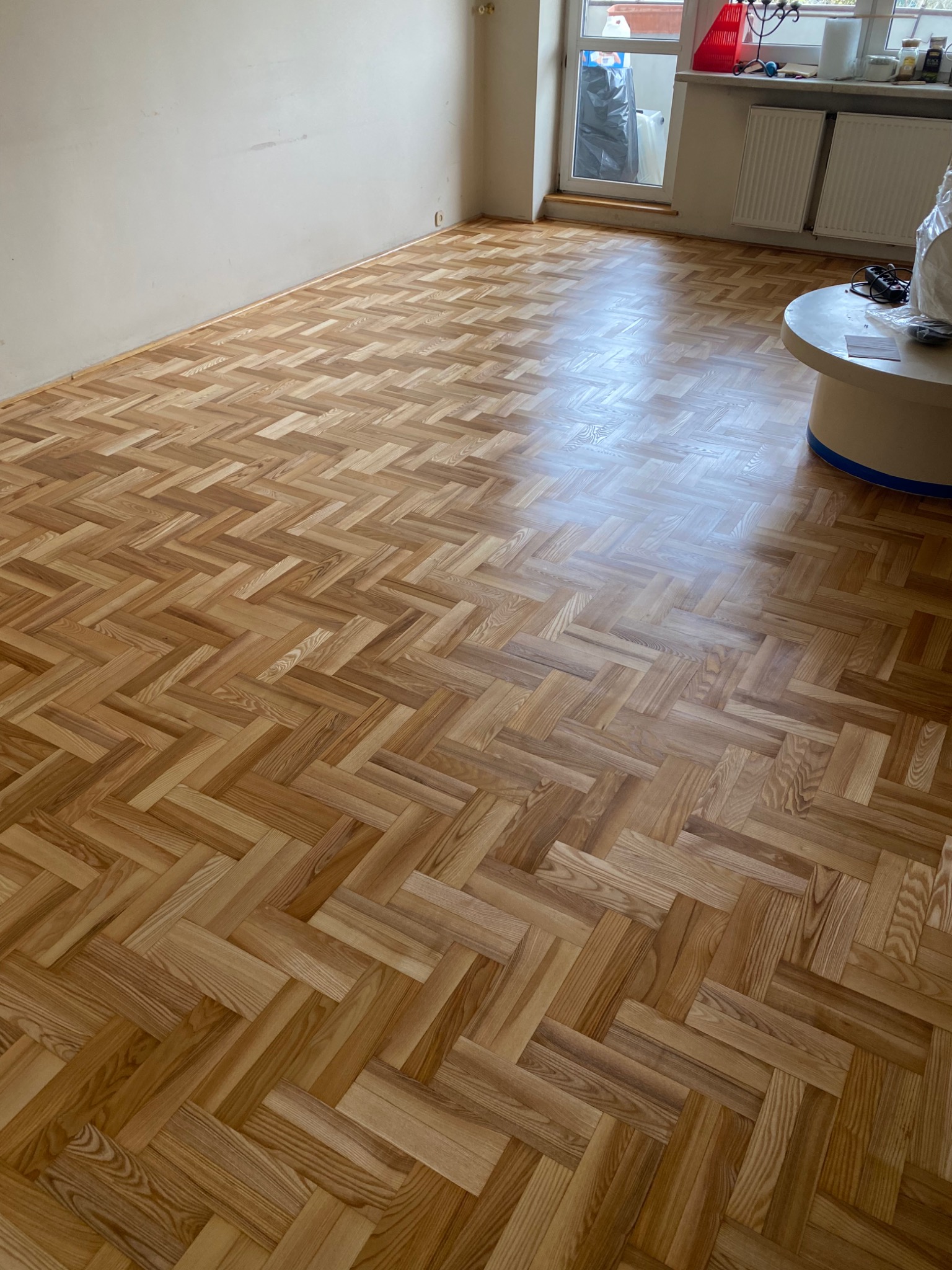 Ukończona podłoga z parkietu ułożonego w jodełkę, widoczne ślady po cyklinowaniu, jasne drewno, w tle okno z grzejnikami i stolik kawowy.
