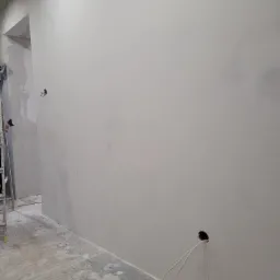 Świeżo otynkowana, gładka ściana wewnętrzna w szarym odcieniu, z wystającymi przewodami elektrycznymi z dwóch otworów w ścianie, podłoga zakurzona gipsem, drabina w tle.