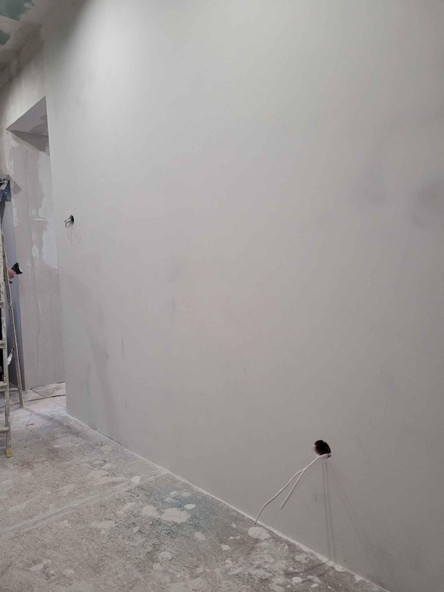 Świeżo otynkowana, gładka ściana wewnętrzna w szarym odcieniu, z wystającymi przewodami elektrycznymi z dwóch otworów w ścianie, podłoga zakurzona gipsem, drabina w tle.