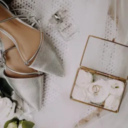 Ułożone srebrne buty na płaskim obcasie z paskami, flakon perfum Versace, pudełko z obrączkami na białych kwiatach i wstążka, tworzące kompozycję ślubną na teksturowanym tle.