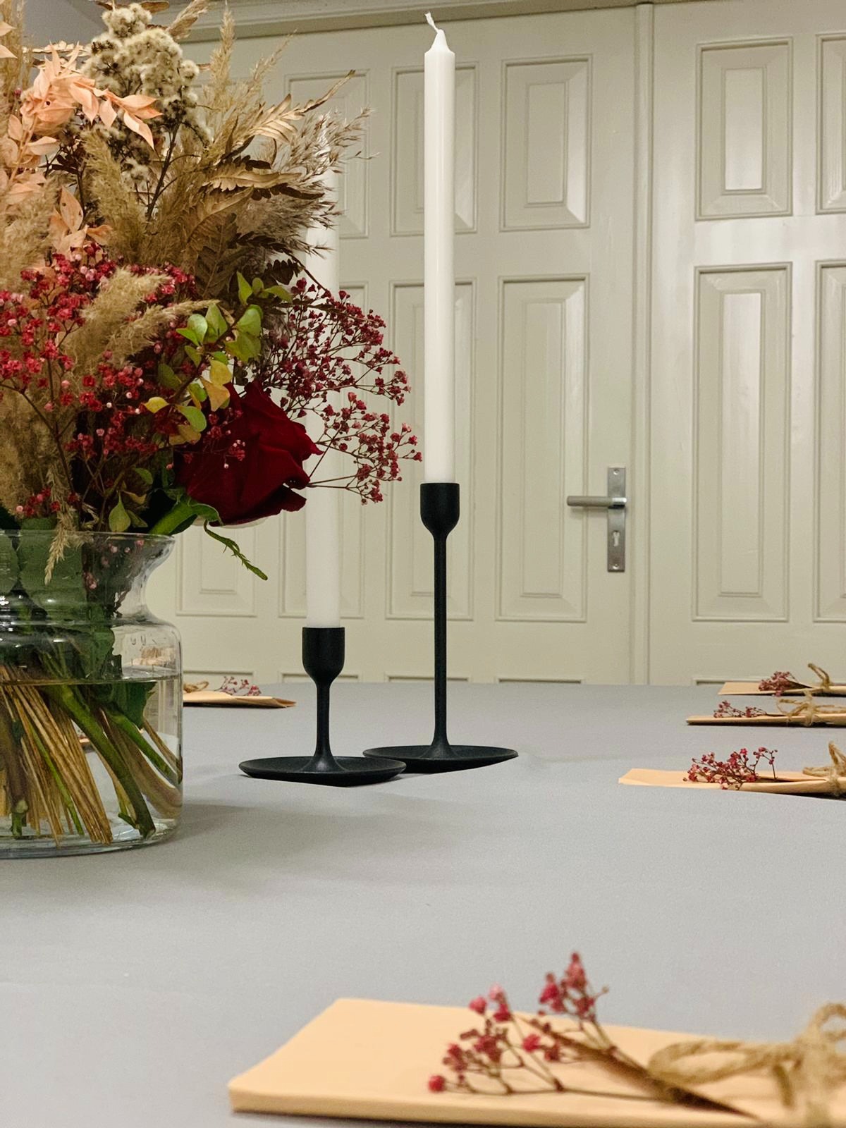 Uroczysta dekoracja stołu z wysokim bukietem z suszonych kwiatów i czerwonych róż, smukłymi białymi świecami w czarnych, minimalistycznych świecznikach oraz drobnymi kompozycjami kwiatowymi...