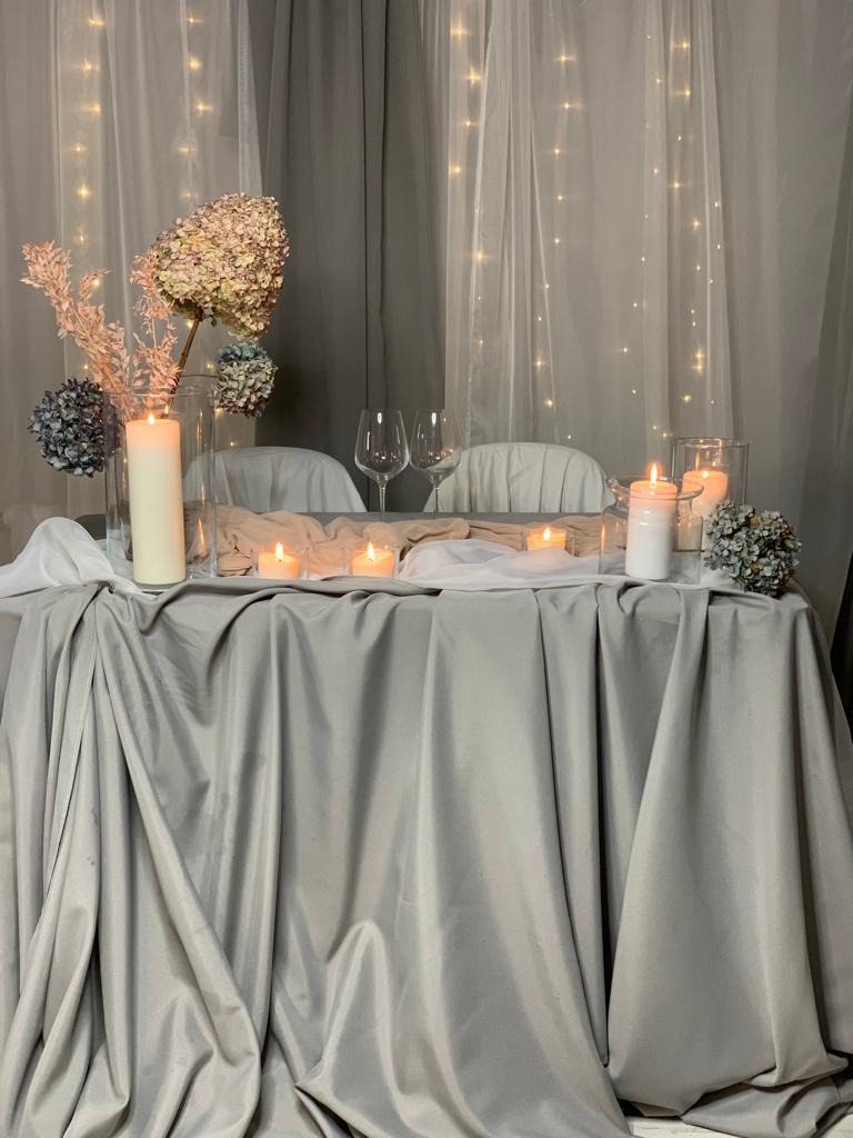 Uroczysta dekoracja stołu z szarą satynową tkaniną, świecami w szklanych naczyniach, kwiatami hortensji i delikatnymi lampkami w tle, tworząca romantyczny nastrój.