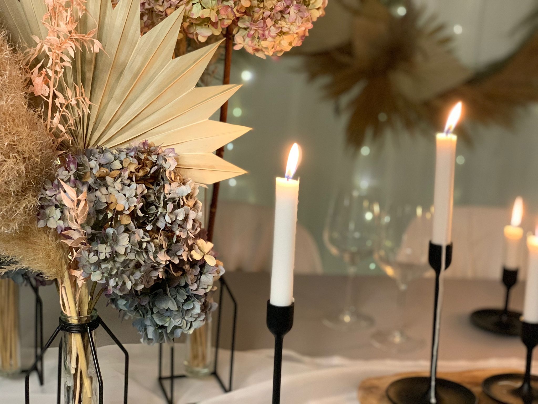 Dekoracja stołu z suszonych kwiatów w szklanych wazonach o metalowej konstrukcji i płonących białych świec w czarnych świecznikach na tle rozmytych światełek.