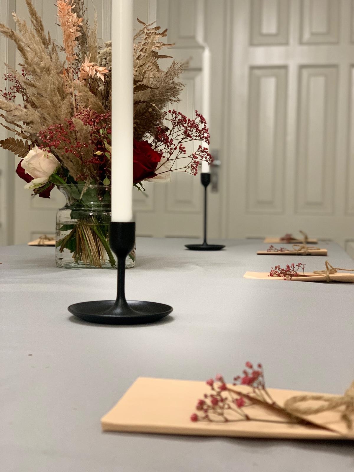 Dekoracja stołu z wysokim bukietem z traw i kwiatów w szklanym wazonie, czarnym świecznikiem i minimalistycznymi wizytówkami z drobnymi czerwonymi kwiatkami, ustawiona na gładkim, szarym stole...