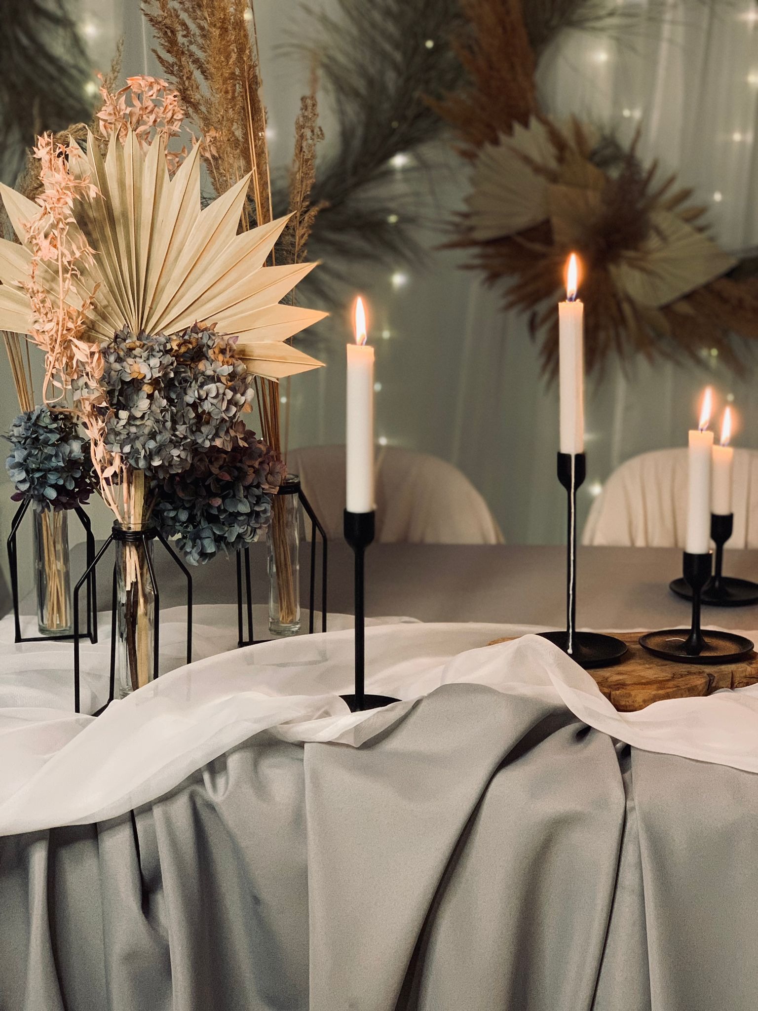 Dekoracja stołu z suszonych kwiatów w geometrycznych wazonach, wysokich czarnych świeczników z płonącymi świecami, całość na szarym obrusie z białym woalem, w tle girlanda świetlna i dekoracja...