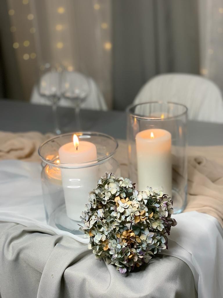 Dekoracja stołu z dwoma świecami w szklanych cylindrach i bukietem suszonych hortensji na tle białych krzeseł i światełek.