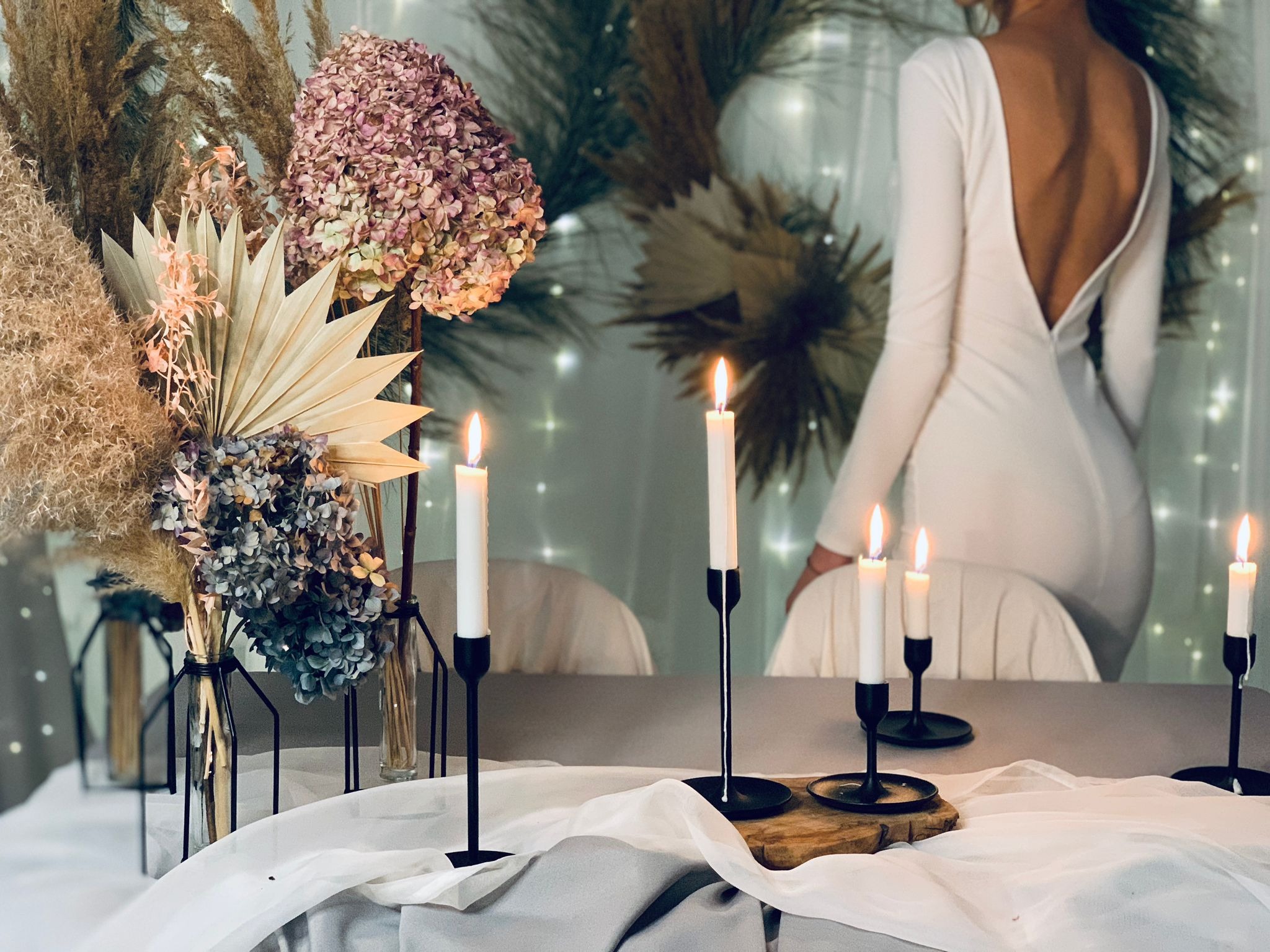 Dekoracja stołu z kompozycjami kwiatowymi w odcieniach beżu i fioletu, wysokimi świecami w czarnych świecznikach, oraz sylwetką kobiety w białej sukni z odkrytymi plecami w tle.