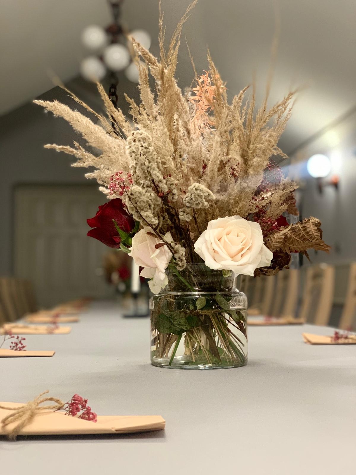 Dekoracja stołu z wysokim bukietem z traw pampasowych, białych i czerwonych róż w szklanym wazonie, ustawiona na szarym obrusie, z widocznymi kartami z podziękowaniami dla gości, przygotowana...