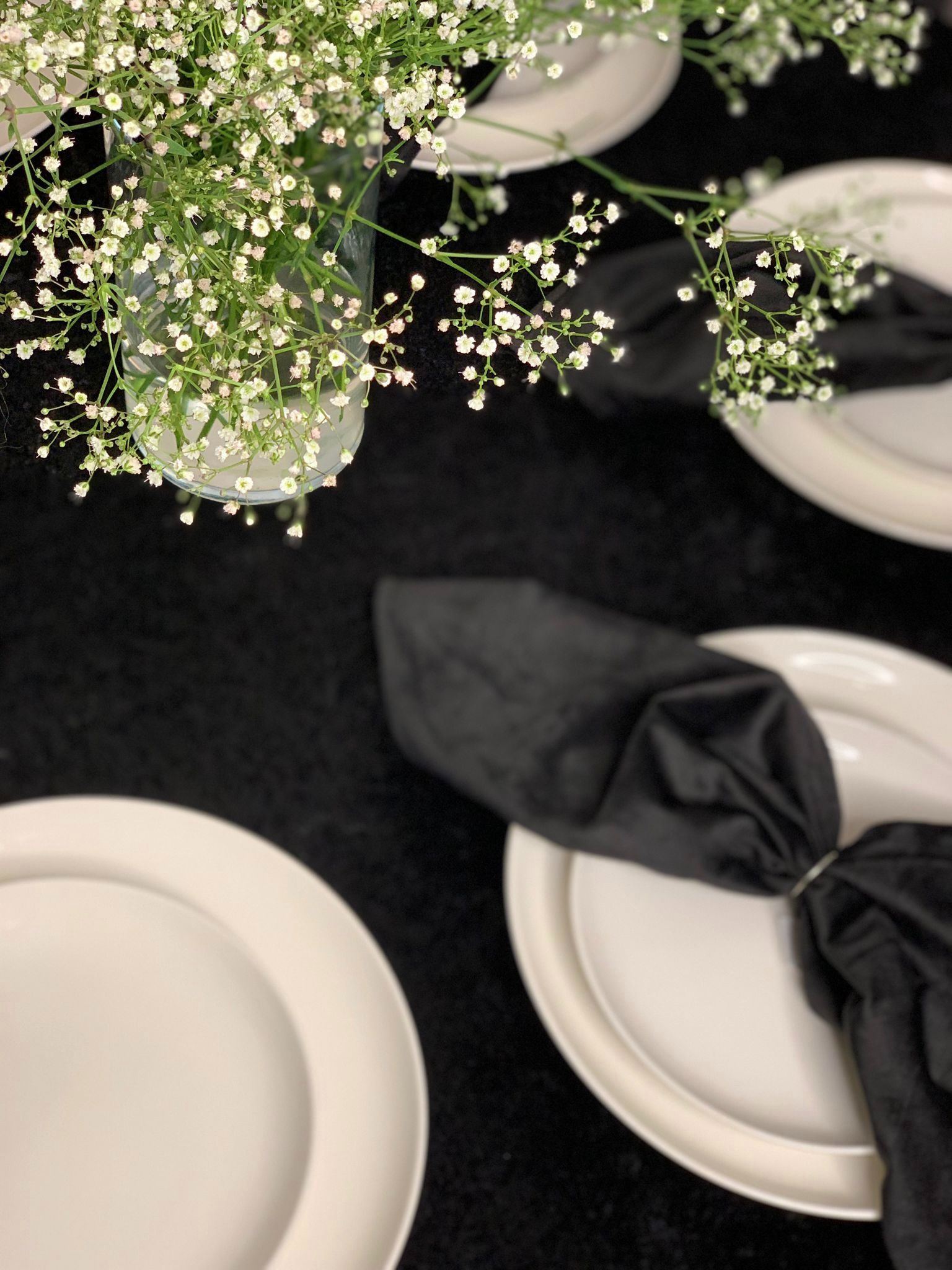 Elegancko nakryty stół z białymi talerzami, czarnymi serwetkami i bukietem gipsówki w szklanym wazonie na czarnym obrusie.