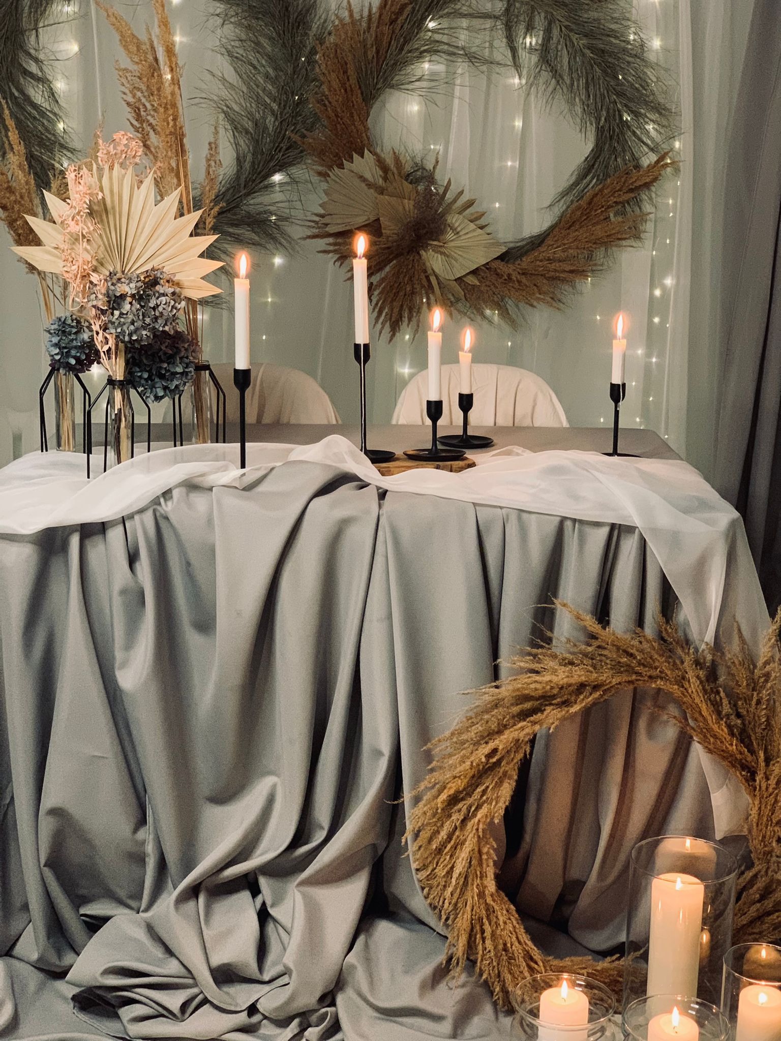 Udekorowany stół z szarą satynową tkaniną, białym woalem, wysokimi czarnymi świecznikami z białymi świecami, kompozycjami z suszonych traw i kwiatów, oraz girlandą z lampek w tle.