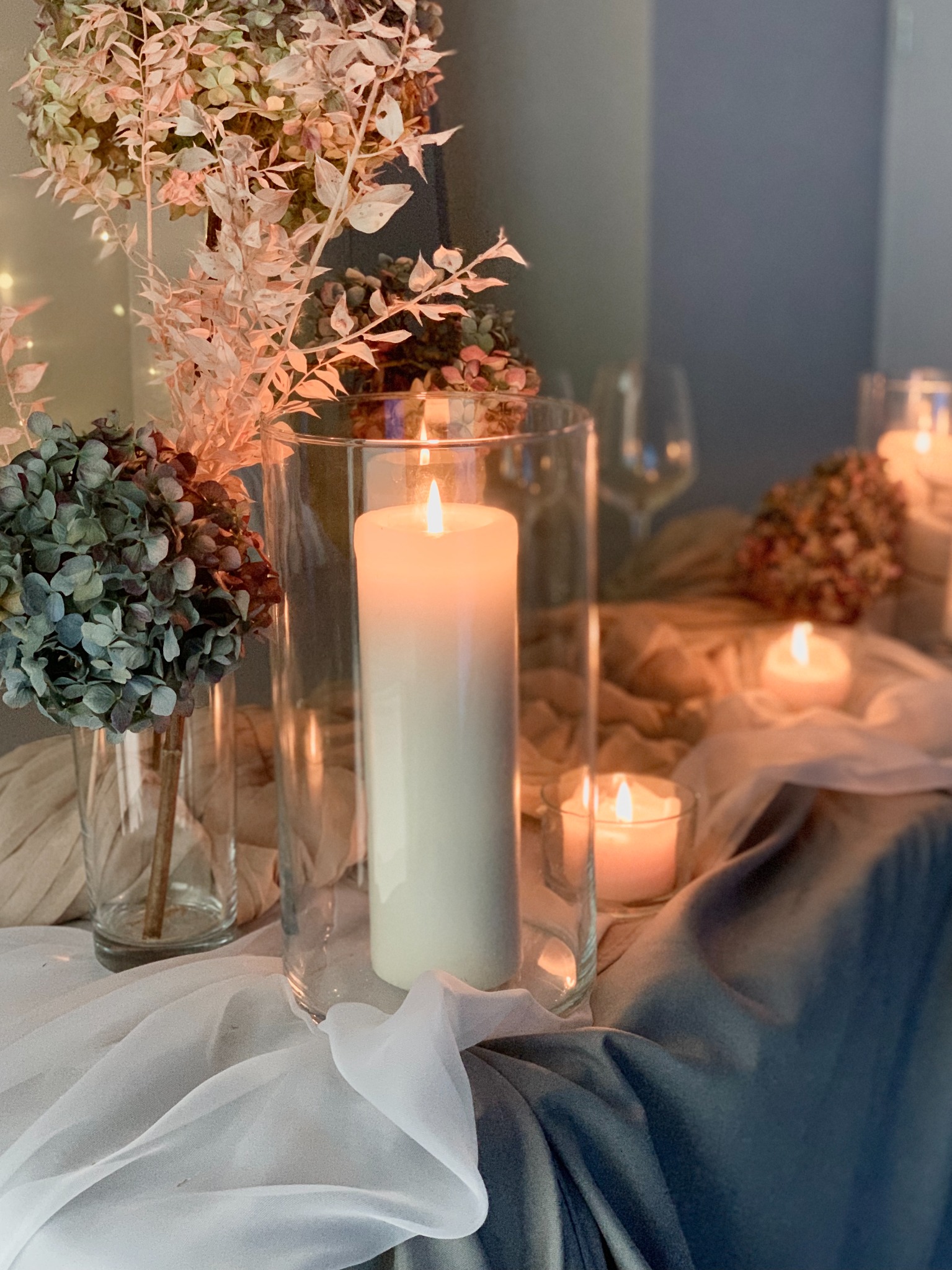Dekoracja stołu z wysokimi świecami w szklanych cylindrach, bukietami hortensji i suszonych kwiatów w wazonach, miękkie światło świec.