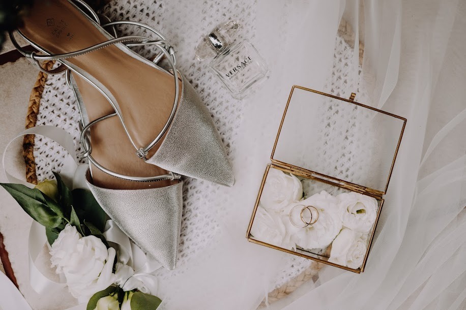 Ułożone srebrne buty na płaskim obcasie z paskami, flakon perfum Versace, pudełko z obrączkami na białych kwiatach i wstążka, tworzące kompozycję ślubną na teksturowanym tle.