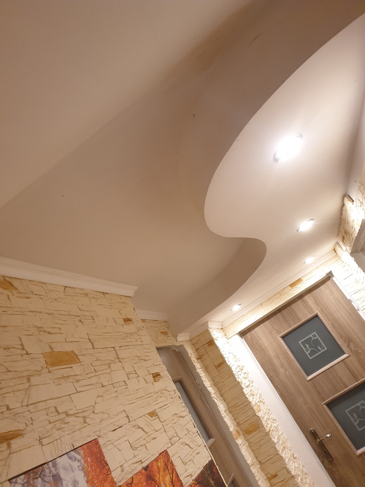 Falowany sufit podwieszany z oświetleniem punktowym, dekoracyjna ściana z imitacji kamienia, drewniane drzwi z przeszkleniami.