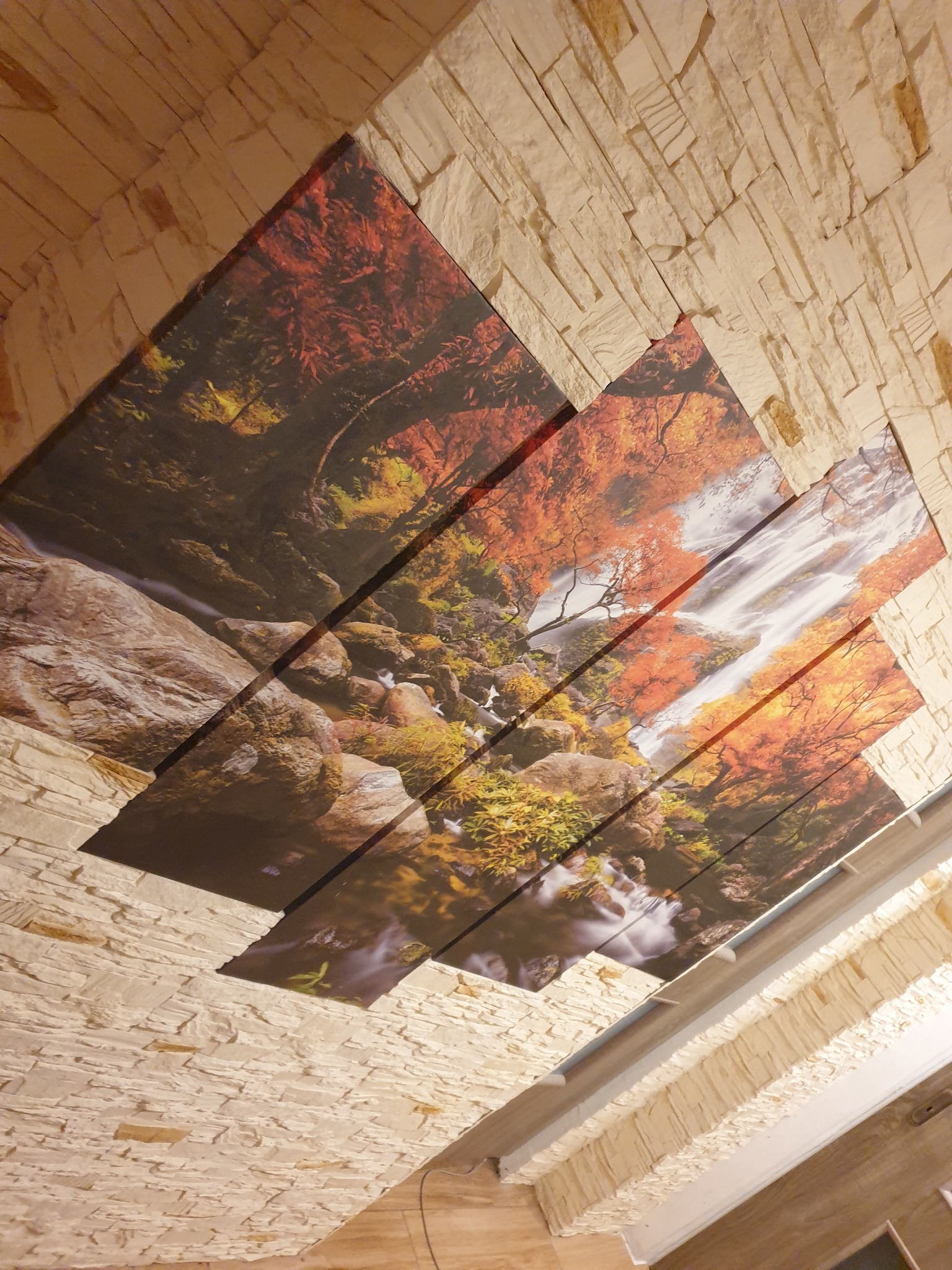 Dekoracyjny sufit z fototapetą przedstawiającą jesienny krajobraz z wodospadem, obramowany imitacją kamiennej okładziny.