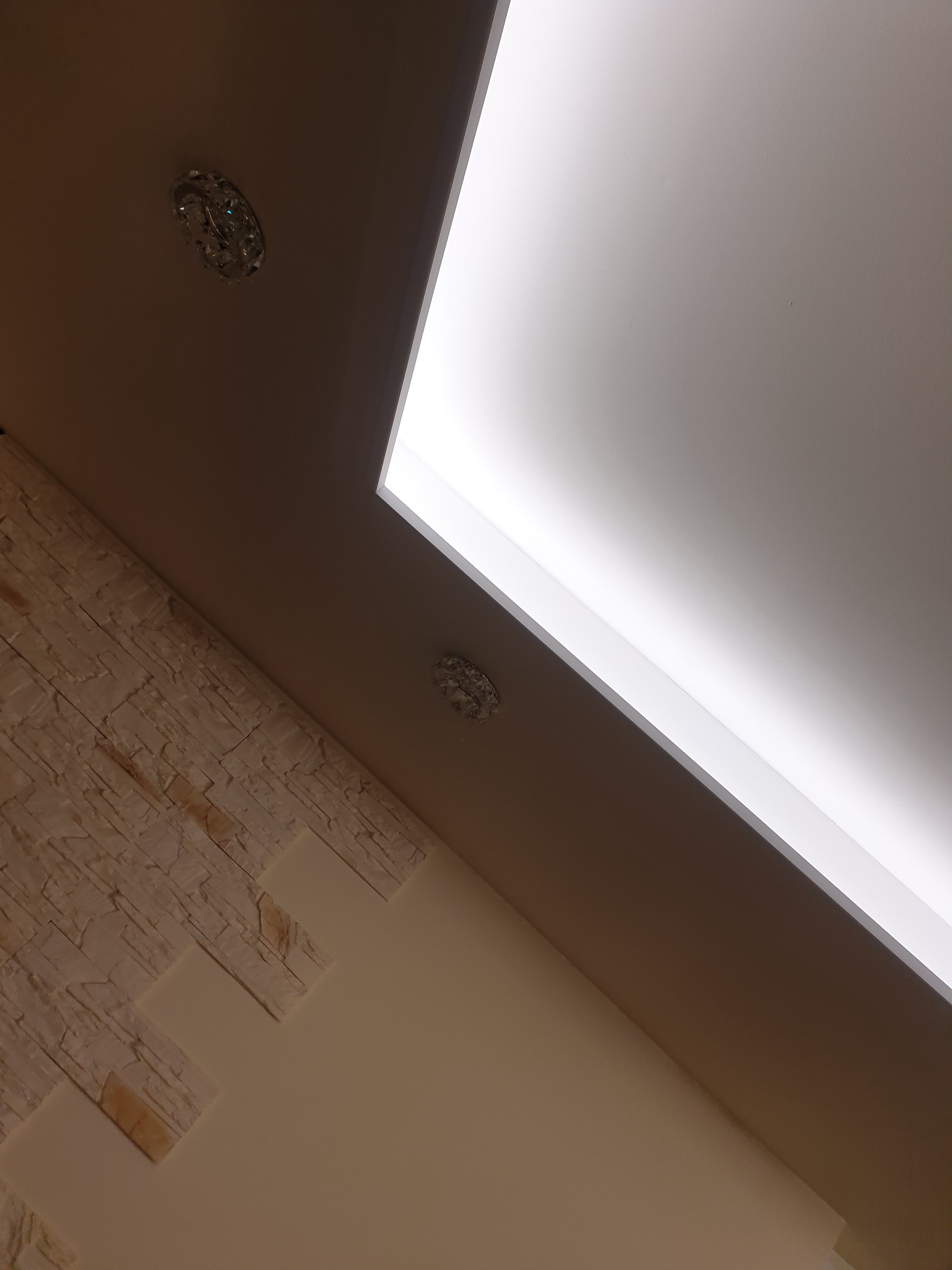 Nowoczesny sufit z podświetleniem LED i ozdobnymi kryształowymi lampkami, połączony z dekoracyjną ścianą wykończoną panelami imitującymi kamień.