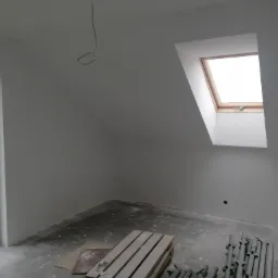 Poddasze po malowaniu na biało, z oknem dachowym, widoczne ślady po remoncie na podłodze, przygotowane do dalszych prac wykończeniowych.