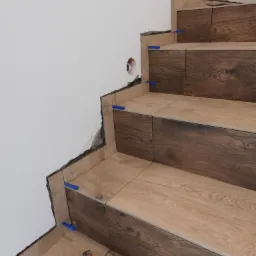 Układanie płytek imitujących drewno na schodach, widoczne niebieskie kliny dystansowe i poziomica, ściana w trakcie wykończenia.