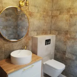 Ukończona łazienka z beżowo-brązowymi płytkami o wzorzystej fakturze, okrągłe lustro w złotej ramie, biała umywalka na drewnianej szafce z złotymi uchwytami, wisząca toaleta, złota lampa ścienna...