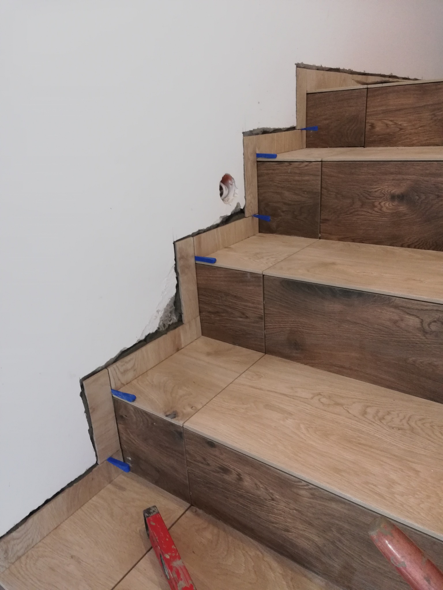 Układanie płytek imitujących drewno na schodach, widoczne niebieskie kliny dystansowe i poziomica, ściana w trakcie wykończenia.