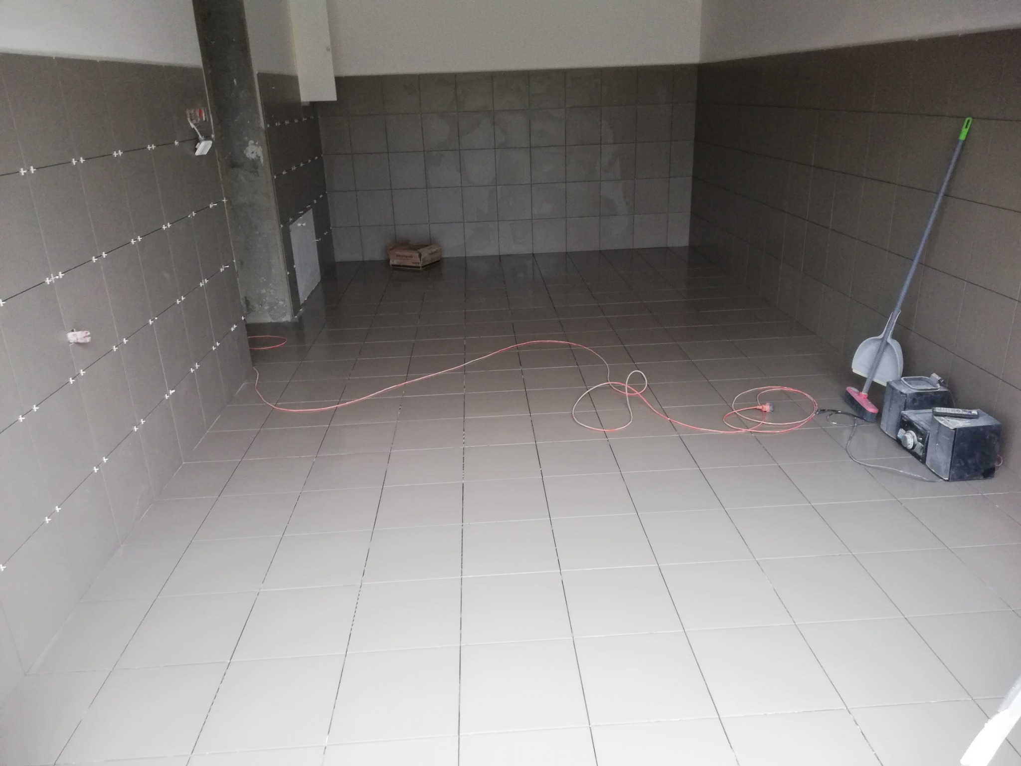 Świeżo wykafelkowane pomieszczenie z jasnoszarymi płytkami, widoczne krzyżyki dystansowe na ścianach, paczka kleju w rogu, kabel zasilający na podłodze oraz sprzęt audio i miotła ze szufelką.