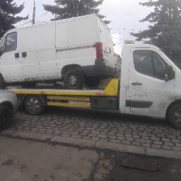 Dominik Grudzień - Biała laweta z białym busem na platformie, widok z boku na tle drzew i chodnika z kostki.