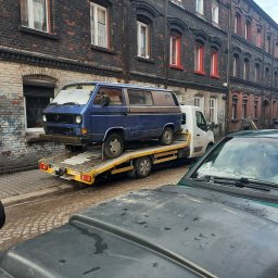 Dominik Grudzień - Niebieski Volkswagen T3 na żółtej lawecie na tle starej kamienicy, częściowo zasłonięty przez zaparkowany samochód z plamami na masce.