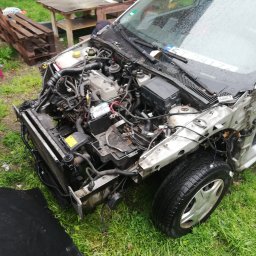 Dominik Grudzień - Uszkodzony przód srebrnego samochodu osobowego z widocznym silnikiem i elementami karoserii, stojący na trawie. Obok widoczny fragment czarnej plandeki i różowe krzesło.