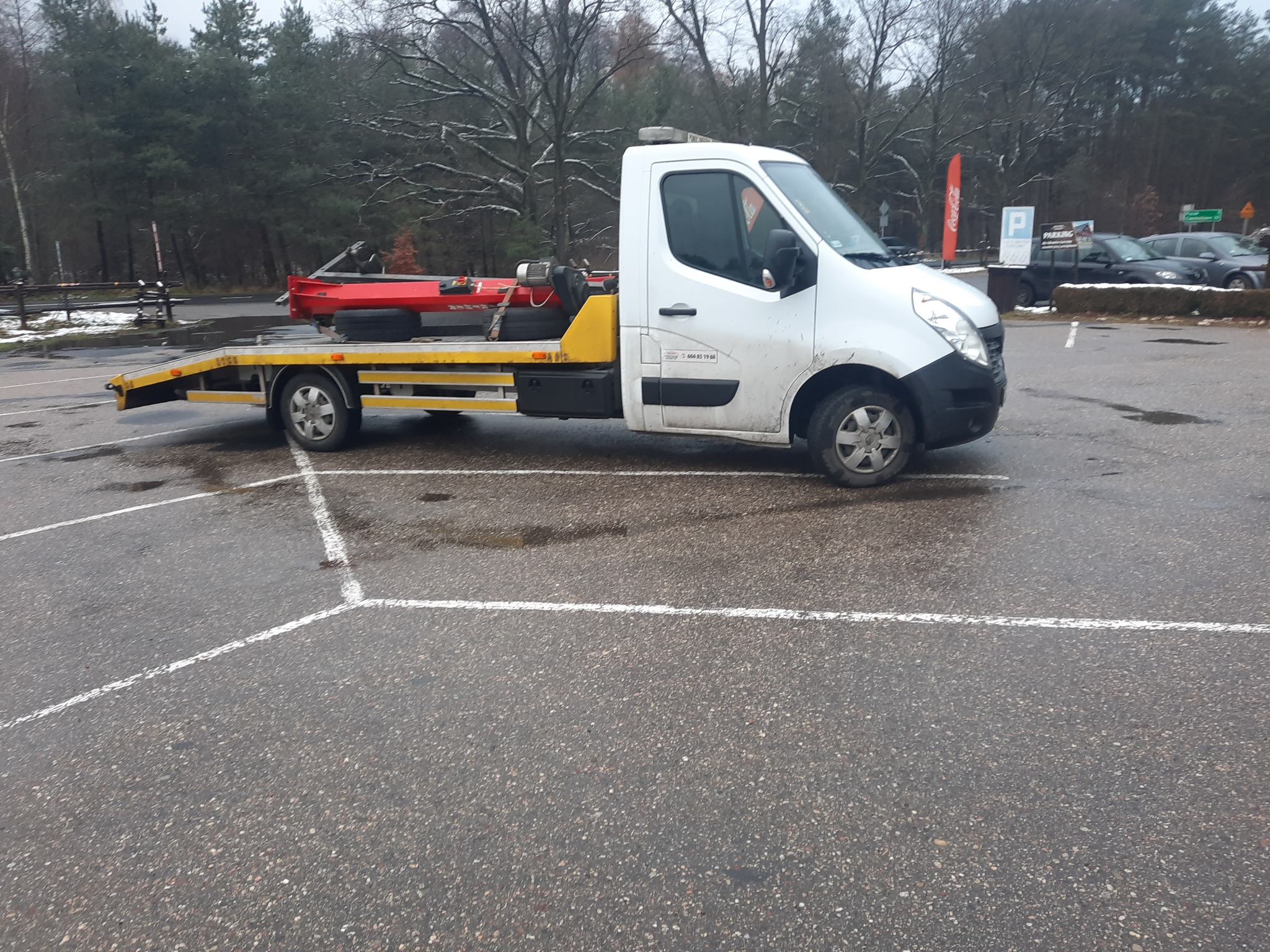 Biała laweta z żółtymi elementami platformy, przewożąca czerwoną konstrukcję, zaparkowana na mokrym parkingu w pobliżu lasu.