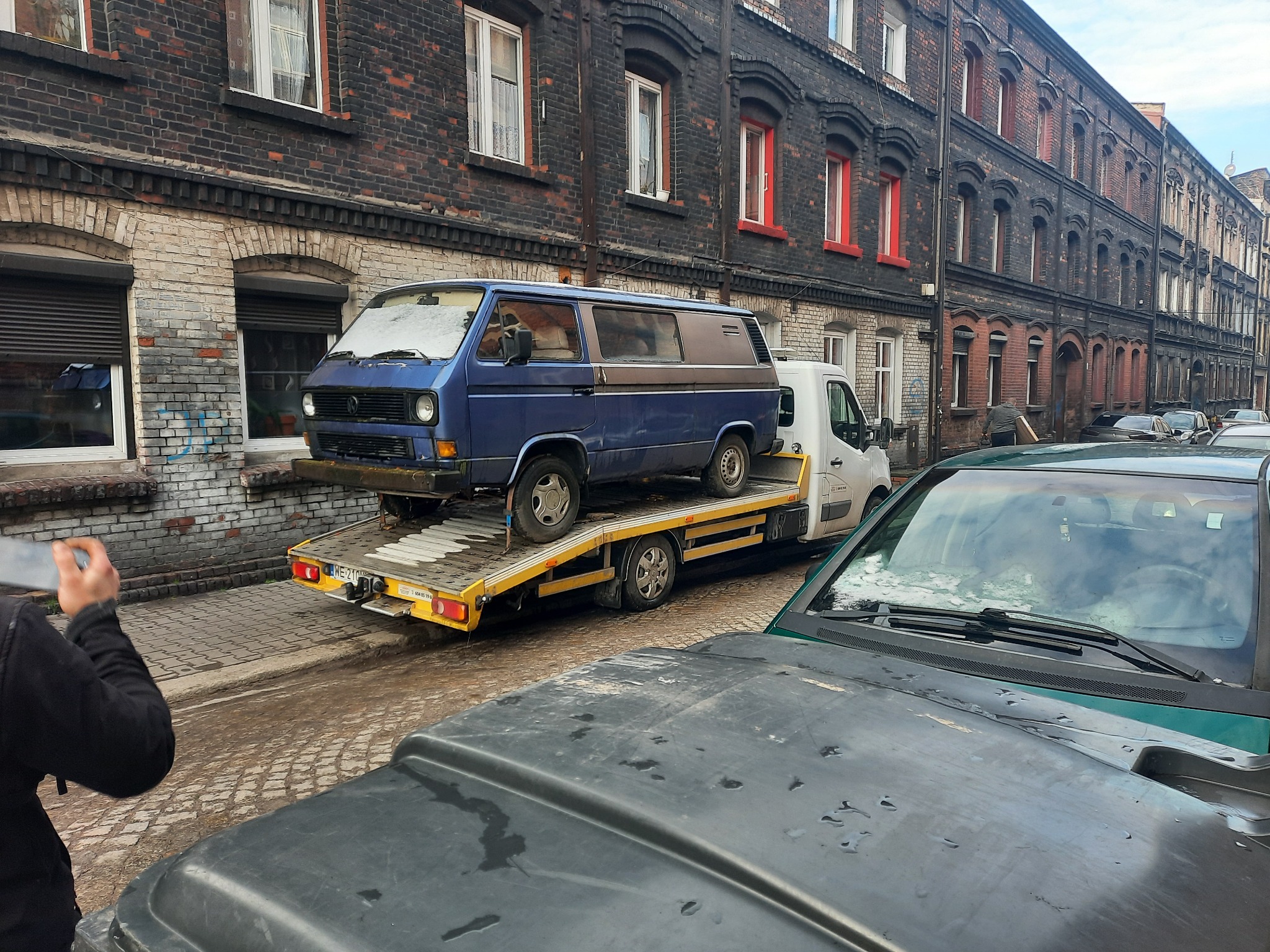 Niebieski Volkswagen T3 na żółtej lawecie na tle starej kamienicy, częściowo zasłonięty przez zaparkowany samochód z plamami na masce.