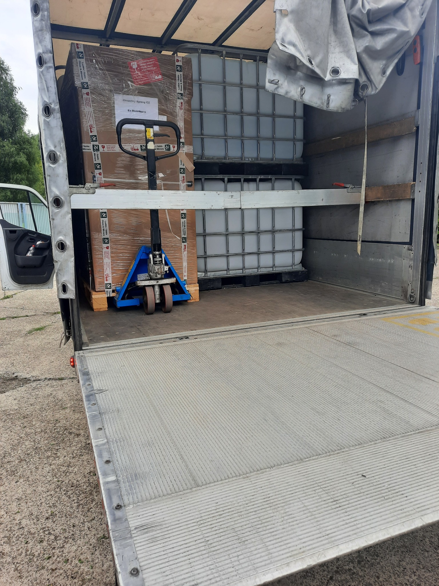 Wnętrze przestrzeni ładunkowej busa z załadowanym towarem na paletach, przesuwane za pomocą wózka paletowego, częściowo zakryte plandeką, widoczne białe pojemniki IBC i zapakowane kartony z napisem...