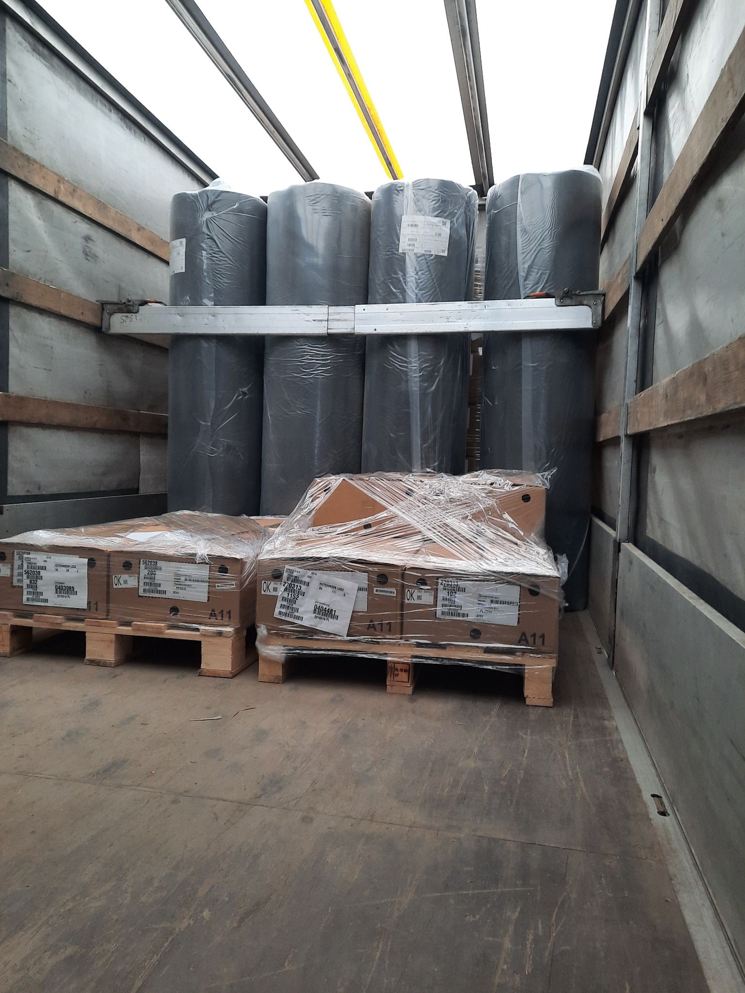 Wnętrze przestrzeni ładunkowej z paletami z kartonami owiniętymi folią oraz pionowo ustawionymi rolkami materiału zabezpieczonymi na metalowych wspornikach.