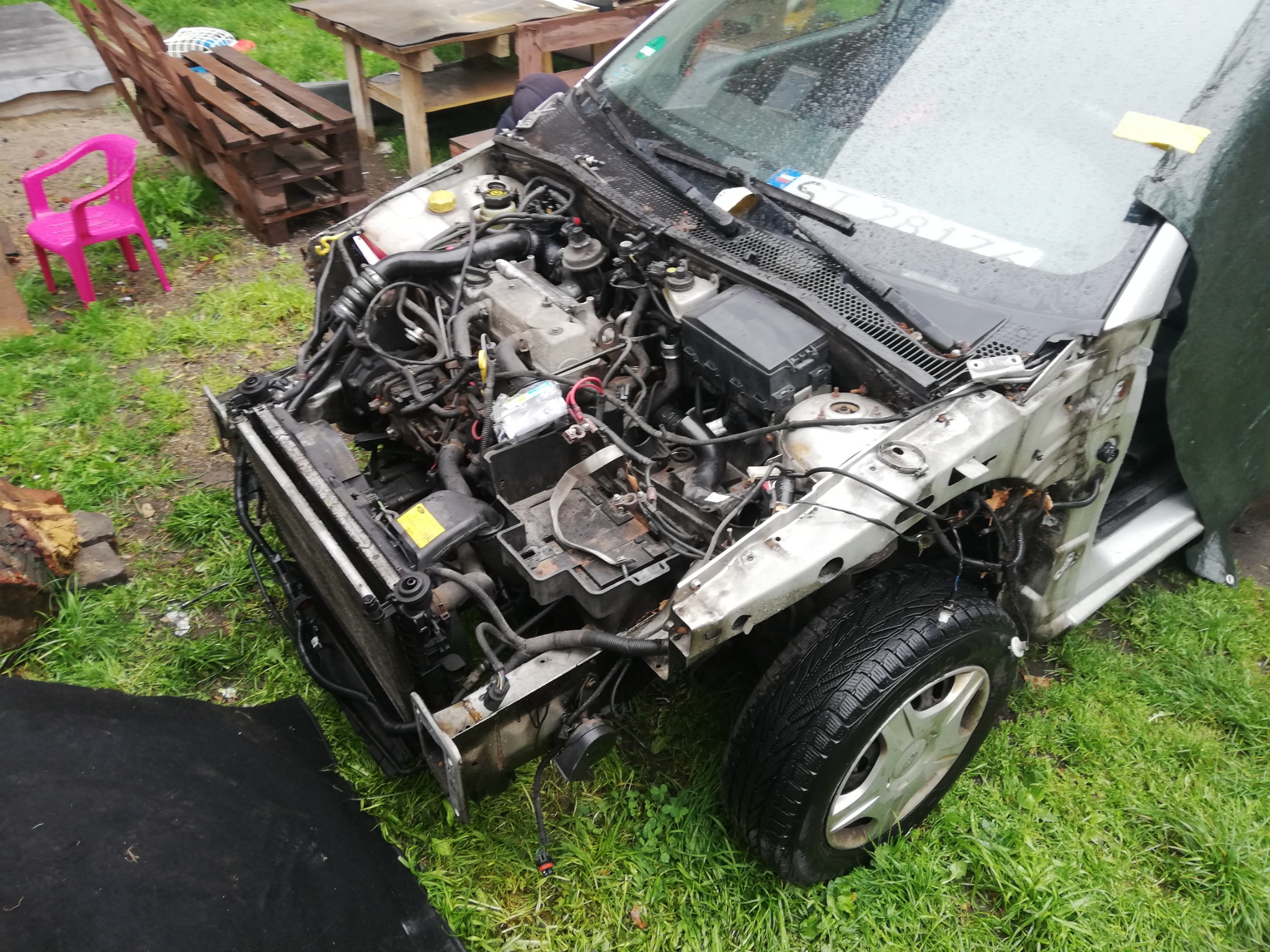 Uszkodzony przód srebrnego samochodu osobowego z widocznym silnikiem i elementami karoserii, stojący na trawie. Obok widoczny fragment czarnej plandeki i różowe krzesło.