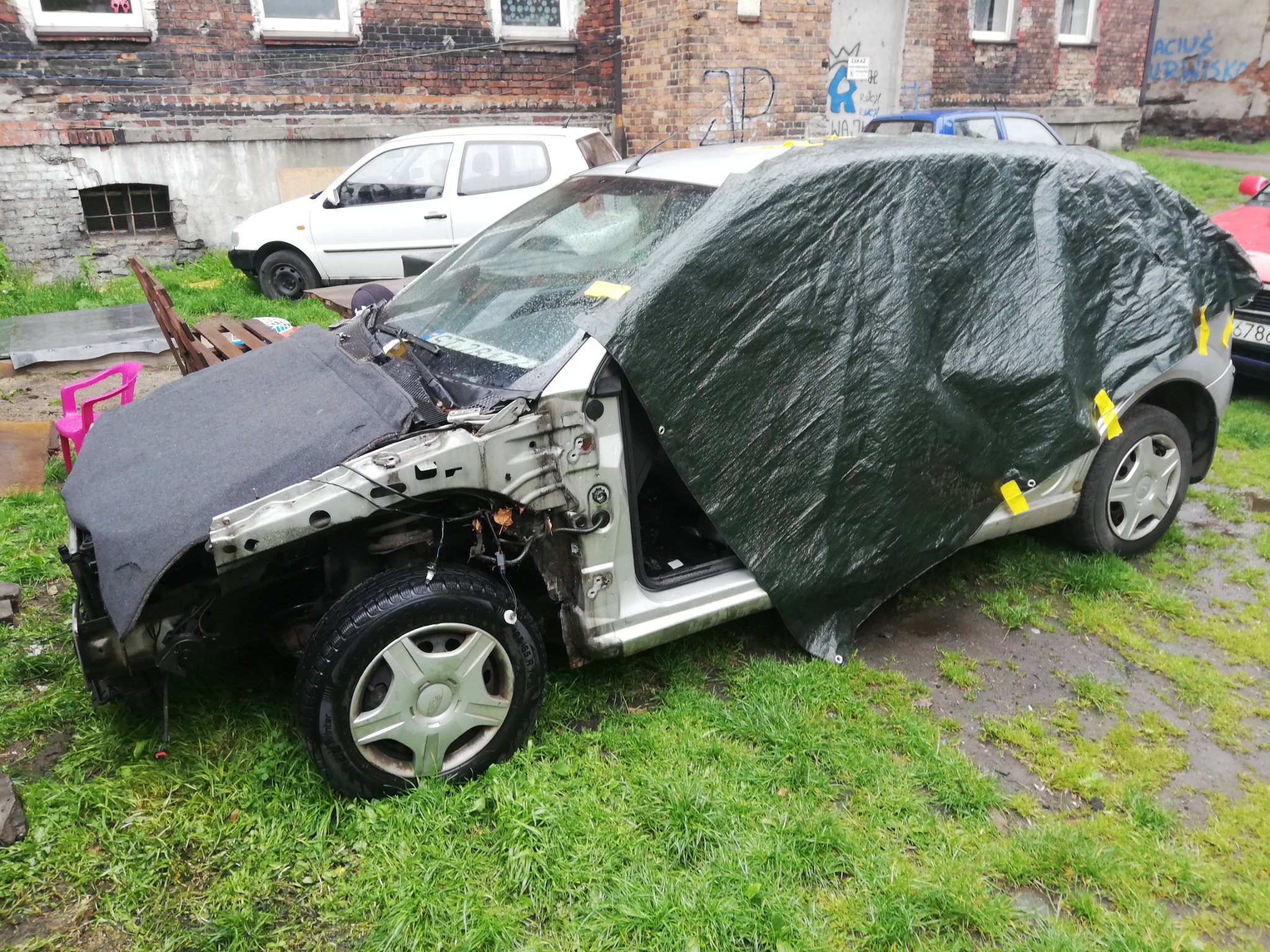 Uszkodzony, srebrny samochód osobowy bez przedniej części karoserii, częściowo przykryty zieloną plandeką, stojący na trawie przed ceglanym budynkiem.