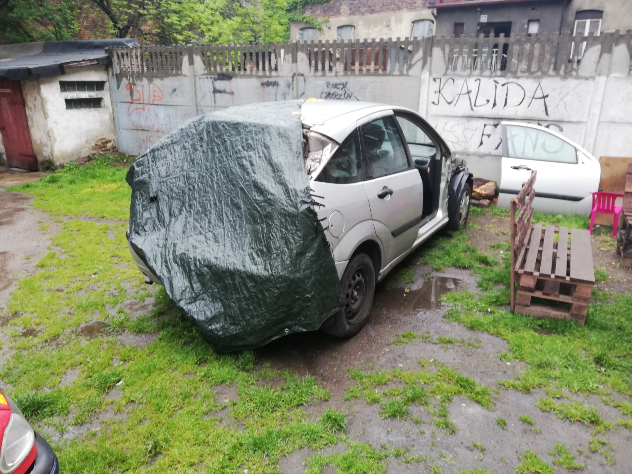 Uszkodzony srebrny samochód osobowy, częściowo przykryty zieloną plandeką, stoi na zewnątrz na trawie, z widocznym ogrodzeniem z graffiti w tle.