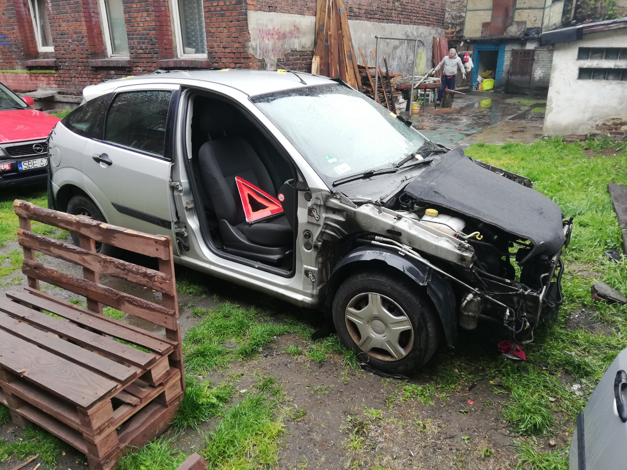 Srebrny samochód po wypadku, z widocznymi uszkodzeniami przodu i otwartymi drzwiami, trójkąt ostrzegawczy wewnątrz, stoi na trawie w zaniedbanym otoczeniu z ceglanym budynkiem w tle.
