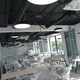 Wnętrze komercyjne w trakcie wykańczania, z widocznymi instalacjami pod sufitem, okrągłymi lampami wiszącymi i meblami zabezpieczonymi folią ochronną.