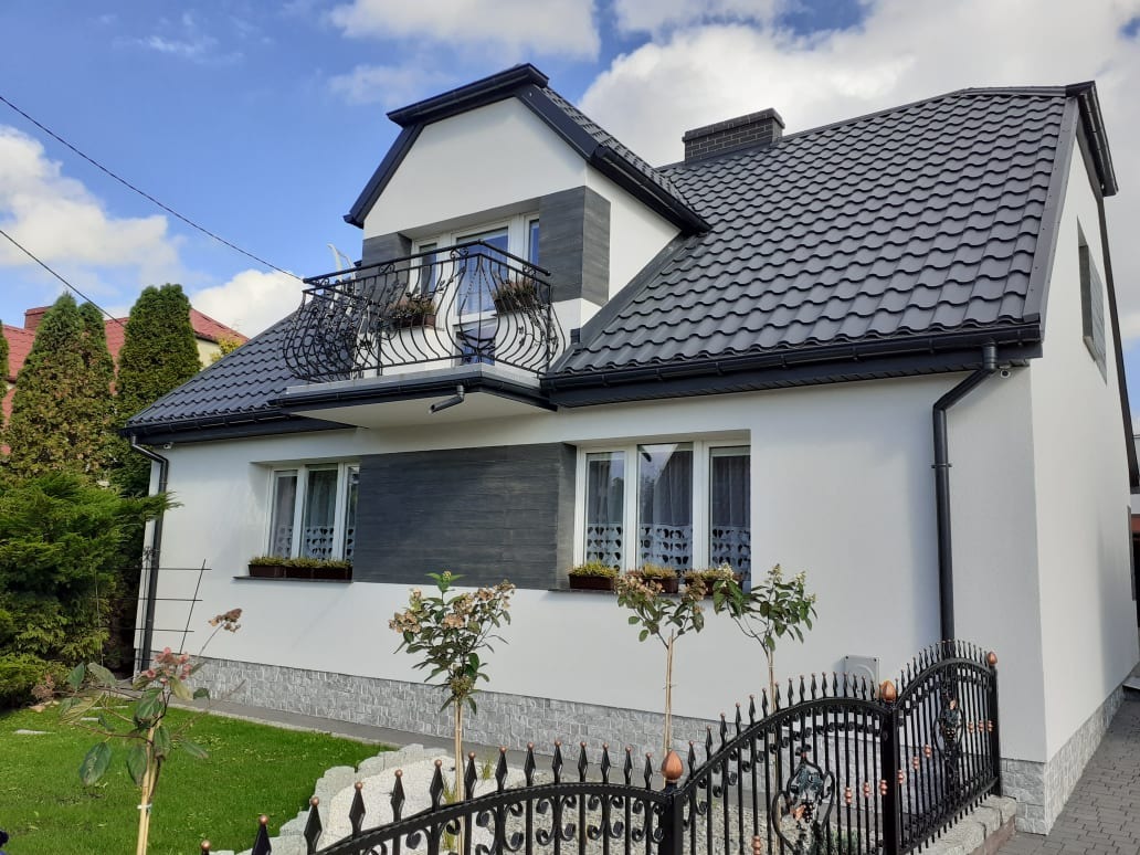 Dwupiętrowy dom z ciemnoszarą dachówką i balkonem z ozdobną, kutą balustradą, biała elewacja z szarymi akcentami i kamienną podmurówką, widoczne rośliny w donicach pod oknami i ozdobne ogrodzenie.
