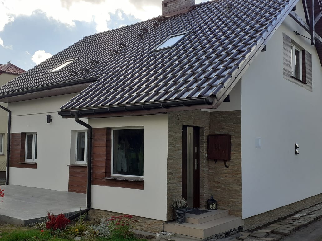 Nowoczesny dom jednorodzinny z ciemnobrązową dachówką, jasną elewacją, fragmentami wykończonymi imitacją drewna i kamienia, z widocznym oknem dachowym i skrzynką na listy.