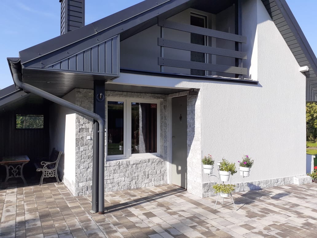 Nowoczesny dom jednorodzinny z elewacją wykończoną szarym kamieniem i białym tynkiem, z czarnym dachem i elementami wykończeniowymi, na pierwszym planie donice z kwiatami.