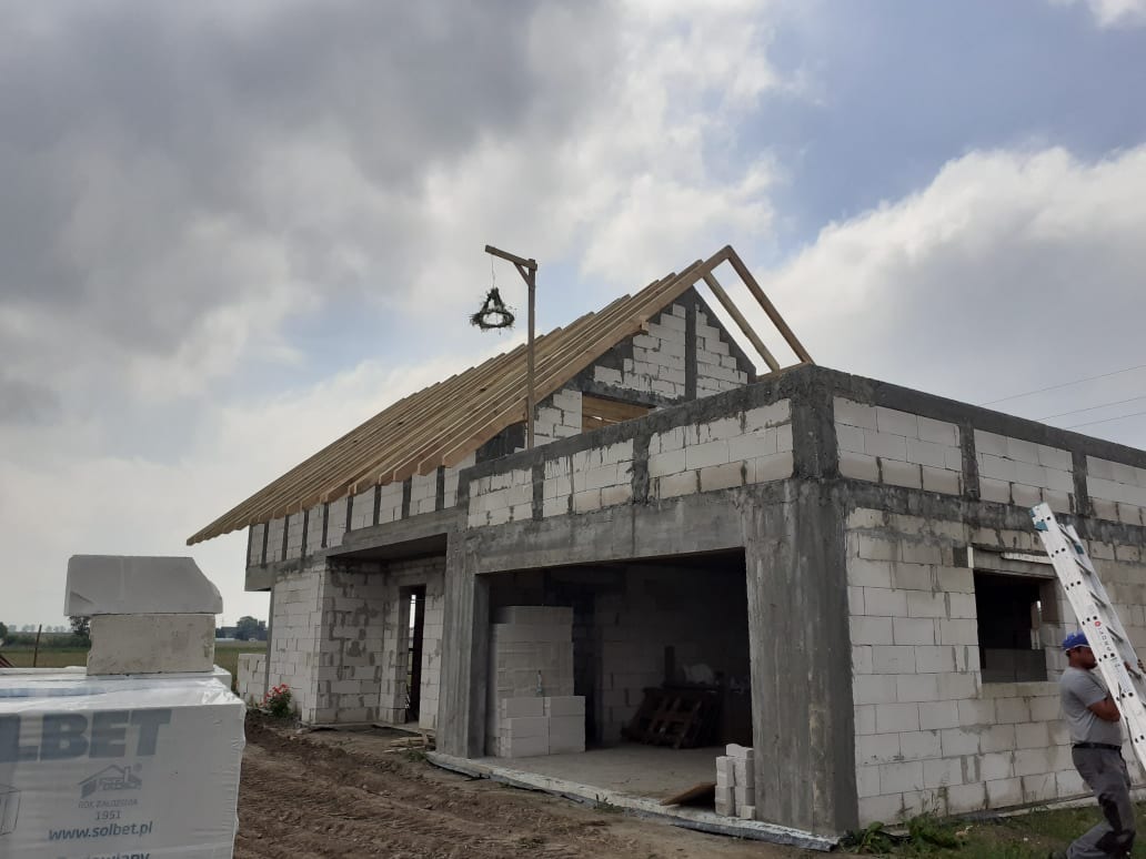 Wznoszenie surowego, jednopiętrowego budynku z białych bloczków, widoczne drewniane elementy konstrukcji dachu, na pierwszym planie paleta z bloczkami Solbet, pracownik oparty o drabinę.