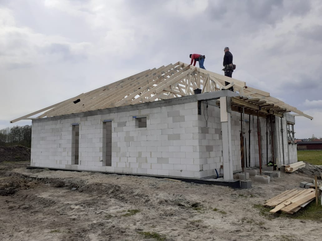 Budowa domu jednorodzinnego z białych bloczków, widoczne drewniane elementy konstrukcji dachu oraz dwóch pracowników na dachu, pochmurne niebo w tle.