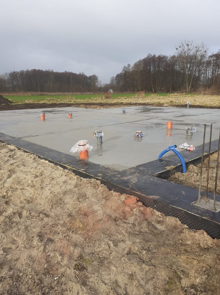 Wylany fundament pod budynek z wystającymi rurami instalacyjnymi zabezpieczonymi folią, widoczna izolacja pionowa fundamentu i stalowe pręty zbrojeniowe, w tle pole i drzewa pod pochmurnym niebem.