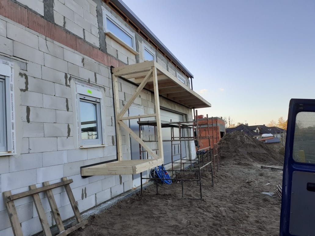 Surowa elewacja domu z betonu komórkowego w trakcie budowy, z widocznym drewnianym rusztowaniem pomocniczym przy oknie i fragmentem otwartego samochodu dostawczego w tle.